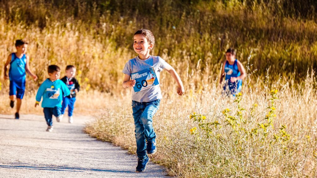 A smiling boy runs on a track through a field while other children run behind him.