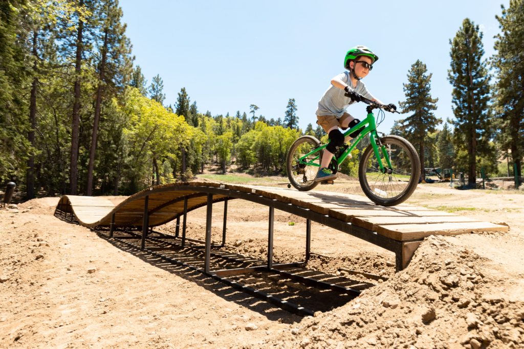 A boy wearing sunglasses, a helmet, and knee pads rides a bike over a wooden bridge on a dirt trail.