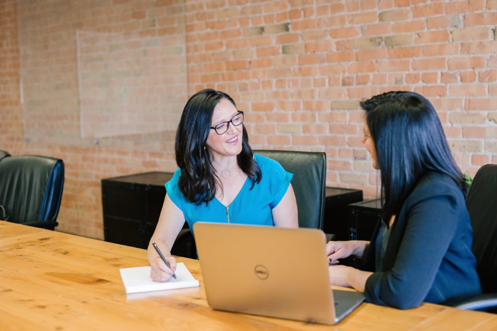 two women sitting at a conference table. one writes on a notepad while the other has a laptop in front of her.