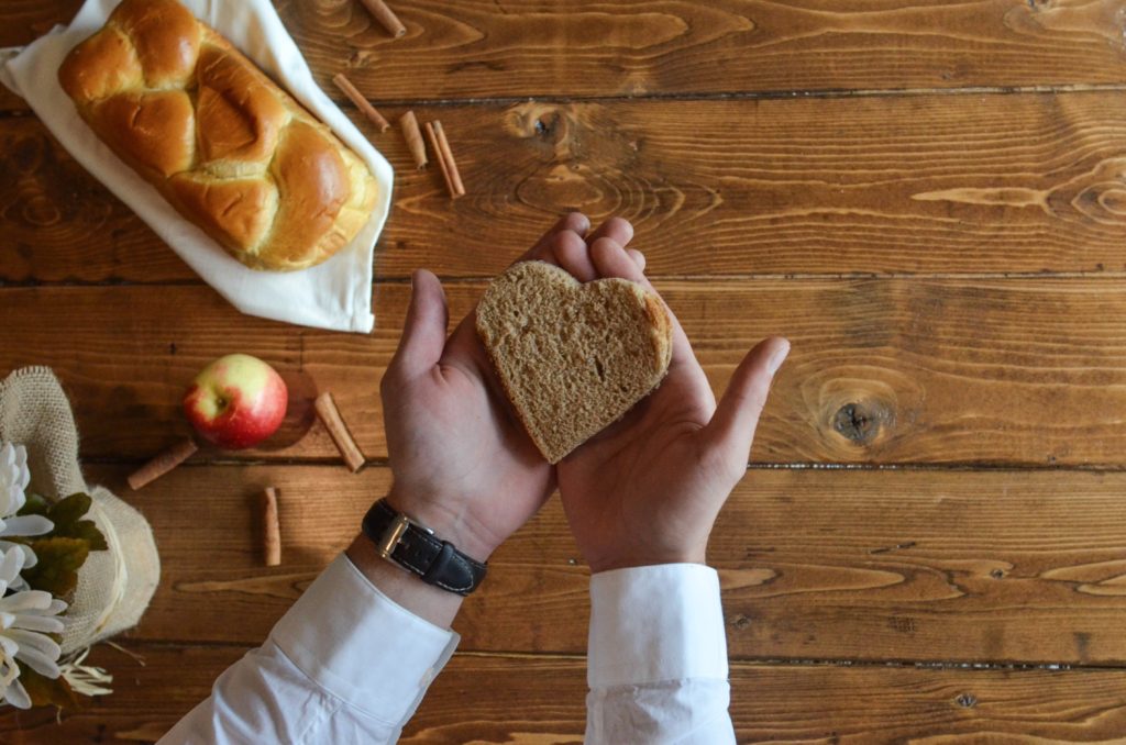 A pair of hands hold a heart-shaped piece of bread over a table with a loaf of bread, an apple, and cinnamon sticks