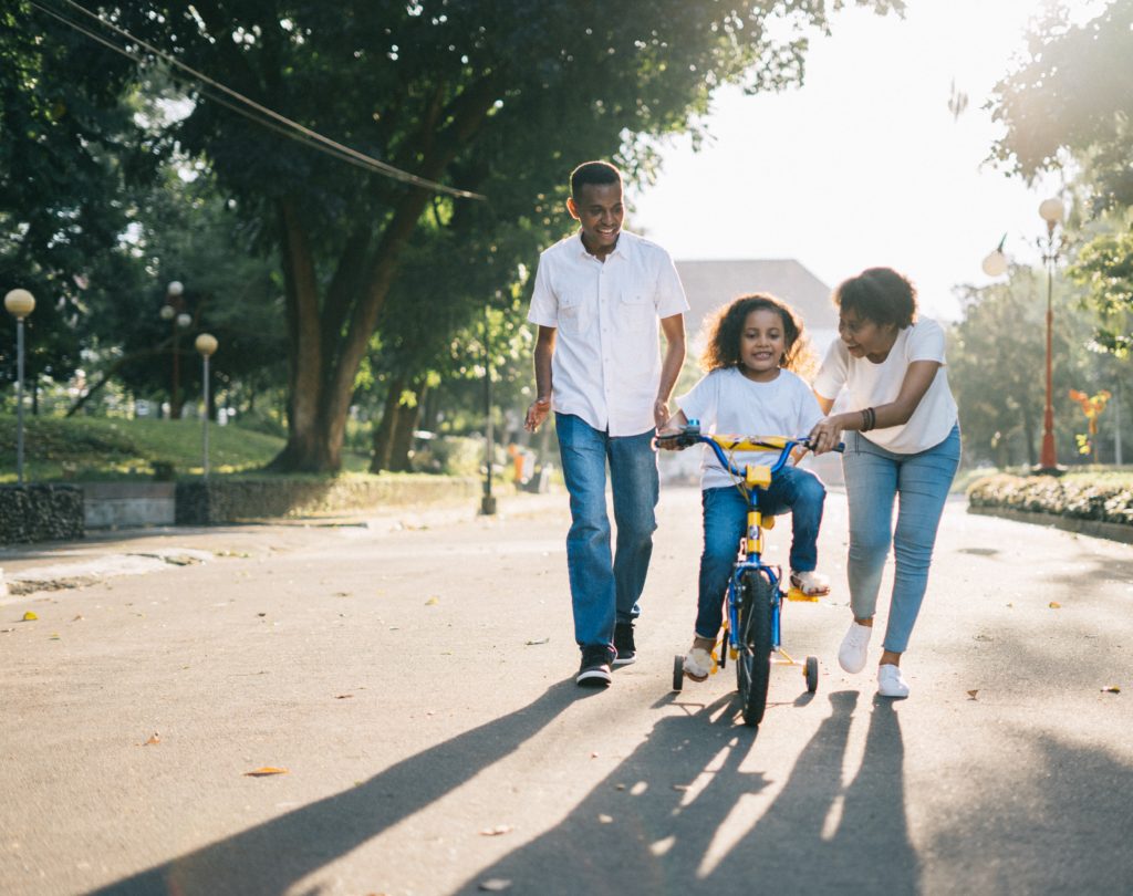 A mother and father helping their daughter ride a bike with training wheels