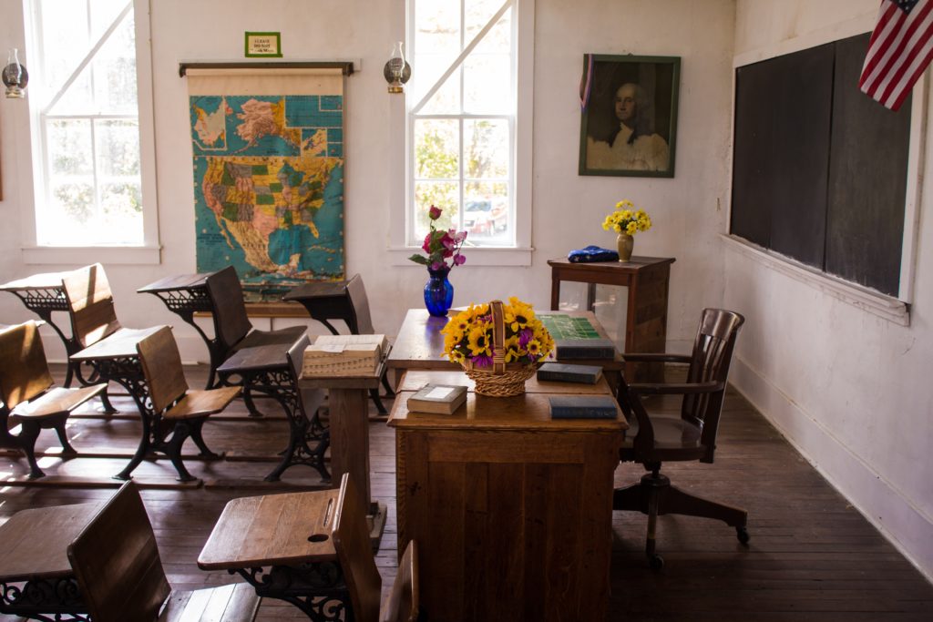 An empty, old-timey schoolhouse with a blackboard on one wall and a map on the back wall between two windows
