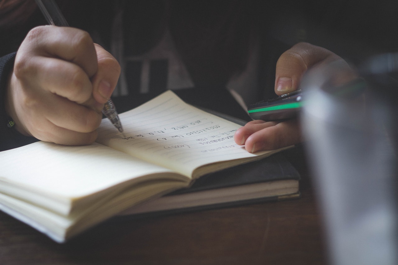 Person writing in notebook while holding calculator