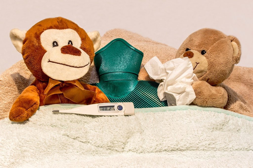Two stuffed animals in bed with a thermometer, hot water bottle, and tissues