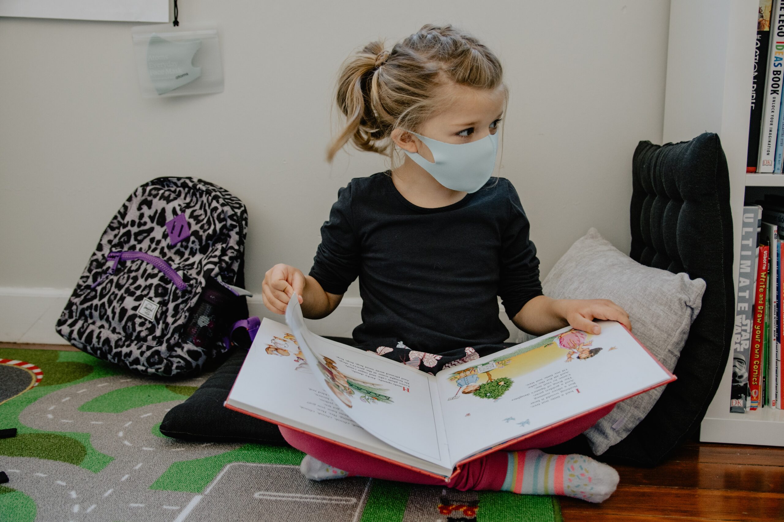 Young student reading book on floor near bookbag