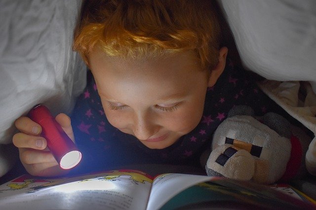 Child reading a book under covers with flashlight